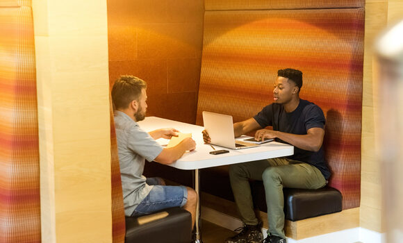 Two young men sat in a cafe booth engaged in conversation while looking at a laptop screen
