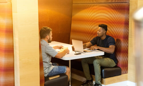 Two young men sat in a cafe booth engaged in conversation while looking at a laptop screen